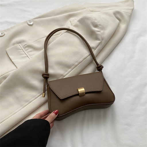 Brown handbag with gold clasp held by a hand on a white surface with a beige coat in the background