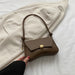 Brown handbag with gold clasp held by a hand on a white surface with a beige coat in the background
