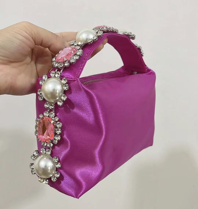 Purple handbag with decorative handle featuring pearls and pink stones held by a hand against a plain background.