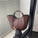 A brown leather tote bag with double top handles, held by a hand in front of a window with white blinds.