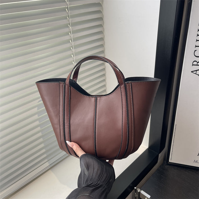 A brown leather tote bag with double top handles, held by a hand in front of a window with white blinds.