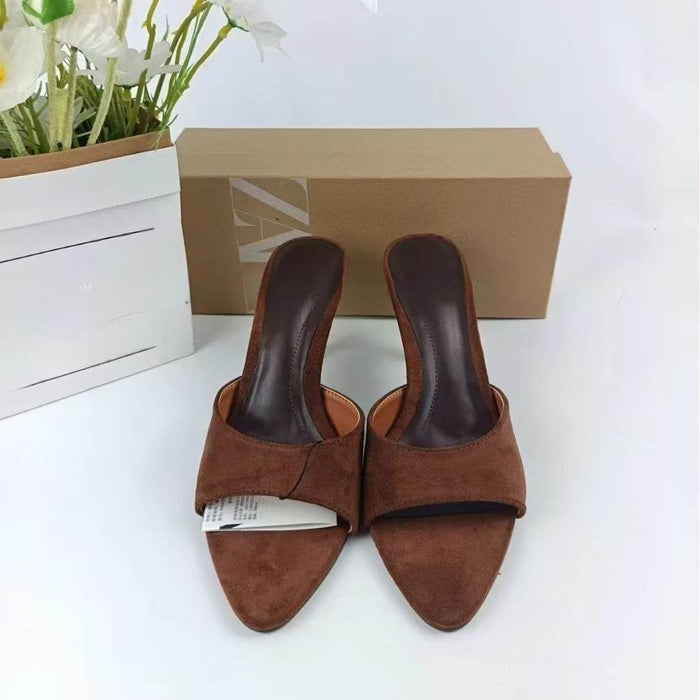 Brown suede mules with a box and flowers in the background on a white surface