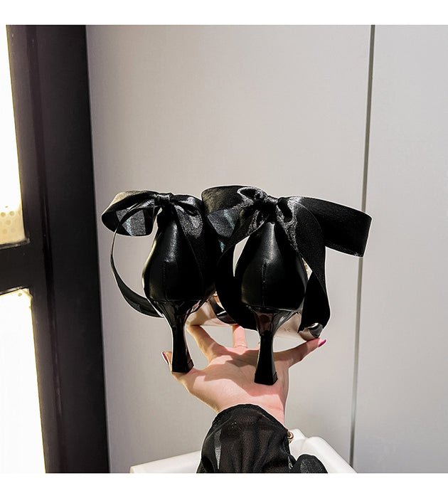 Black high-heeled shoes with large bows held by a person against a neutral background