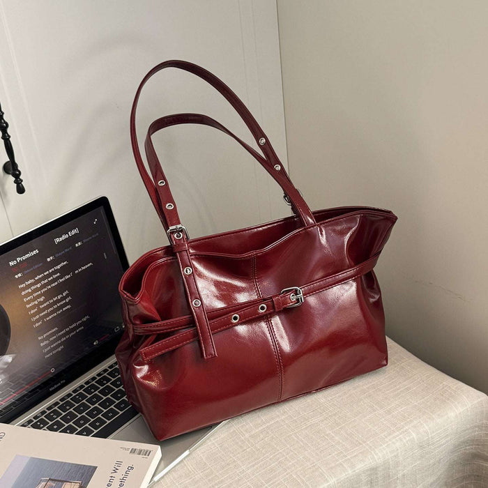 A glossy deep red leather handbag with double shoulder straps and silver-tone hardware, styled on a light-colored fabric-covered surface next to an open laptop.