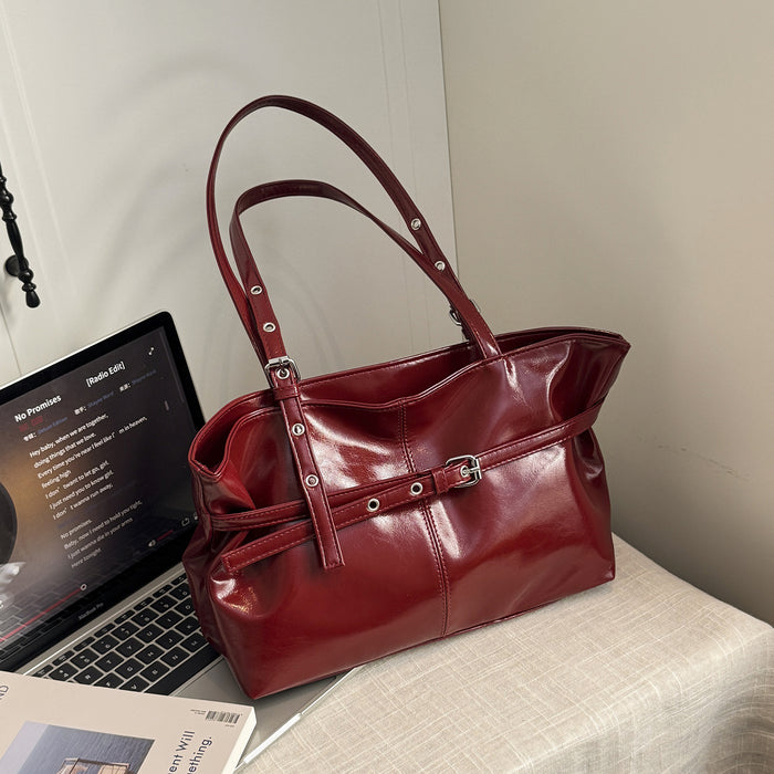 A glossy deep red leather handbag with double shoulder straps and silver-tone hardware, styled on a light-colored fabric-covered surface next to an open laptop.
