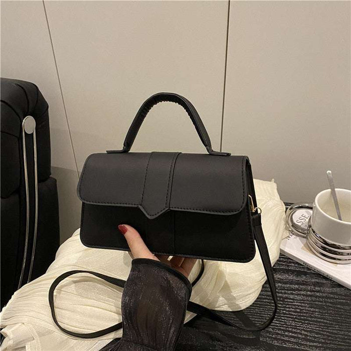 A black faux-leather handbag with a top handle and shoulder strap, held by a hand wearing a sheer black sleeve. The handbag is placed on a white textured blanket with a black suitcase and a white cup in the background.