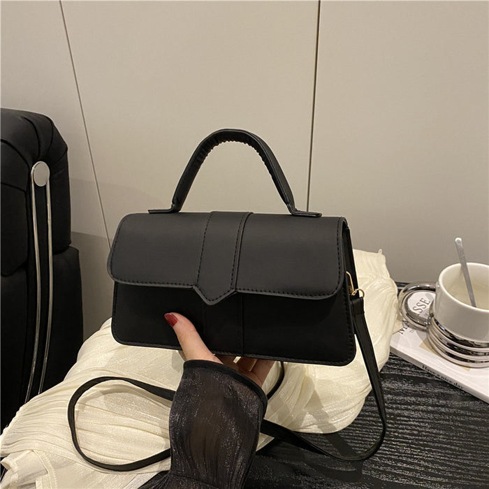 A black faux-leather handbag with a top handle and shoulder strap, held by a hand wearing a sheer black sleeve. The handbag is placed on a white textured blanket with a black suitcase and a white cup in the background.