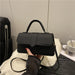 A black faux-leather handbag with a top handle and shoulder strap, held by a hand wearing a sheer black sleeve. The handbag is placed on a white textured blanket with a black suitcase and a white cup in the background.