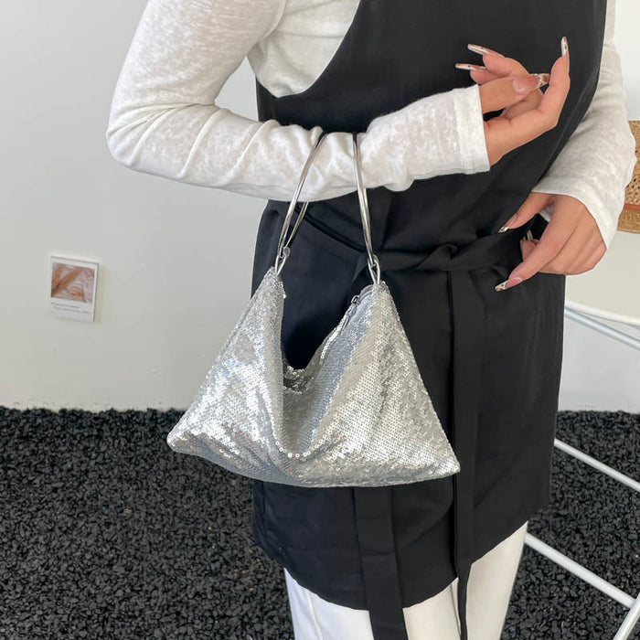 A person wearing a white long-sleeve top and a black apron-style garment holds a silver sequined slouchy shoulder bag with thin metallic handles.