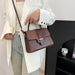 A woman wearing a white blazer and light blue jeans carries a brown leather handbag with a top handle and shoulder strap. The bag features a smooth front panel, a crocodile-embossed flap, and gold-tone hardware.