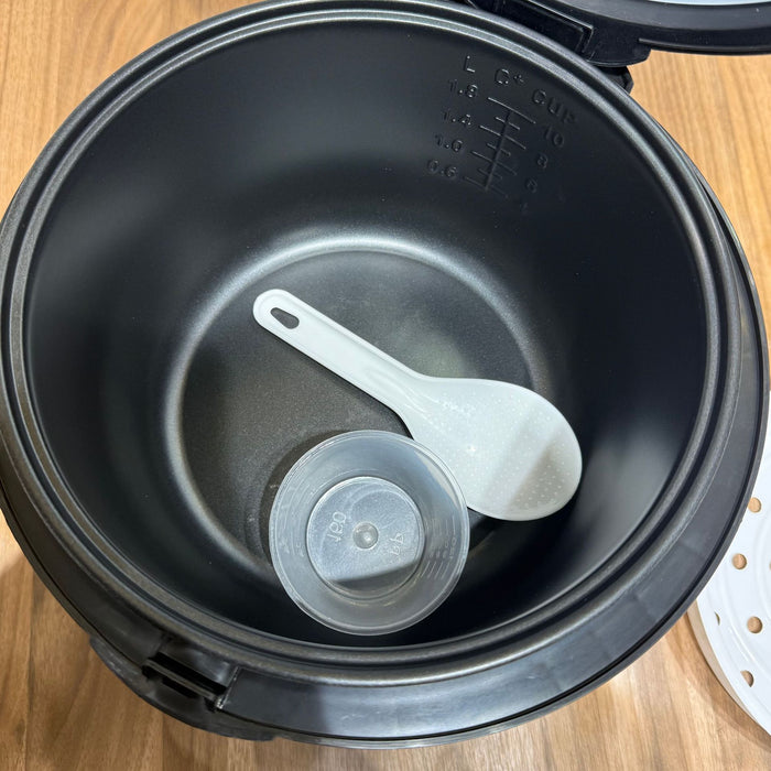 Black rice cooker with a white spoon and clear lid inside on a wooden surface