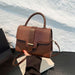 A brown handbag with a mix of smooth and crocodile-embossed textures, held by a hand with red-painted nails over a white textured background.
