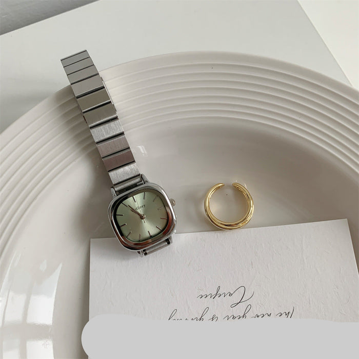 A silver-toned metal watch with a square face and matching metal link bracelet, placed on a white plate next to a gold ring and a white card.