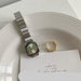 A silver-toned metal watch with a square face and matching metal link bracelet, placed on a white plate next to a gold ring and a white card.