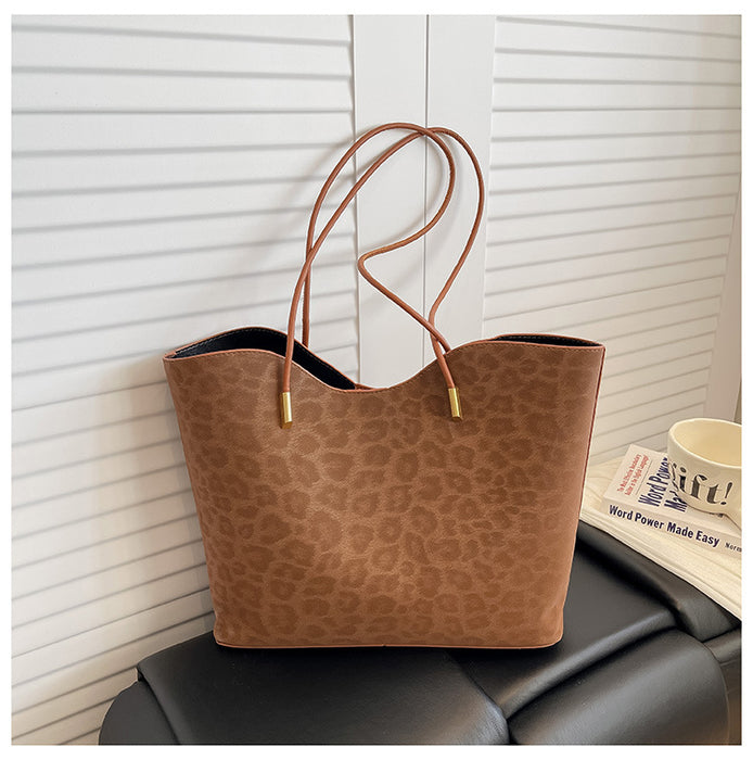 A brown leopard-print tote bag with two long shoulder straps and gold-tone hardware, resting on a black leather sofa in front of white paneled walls.
