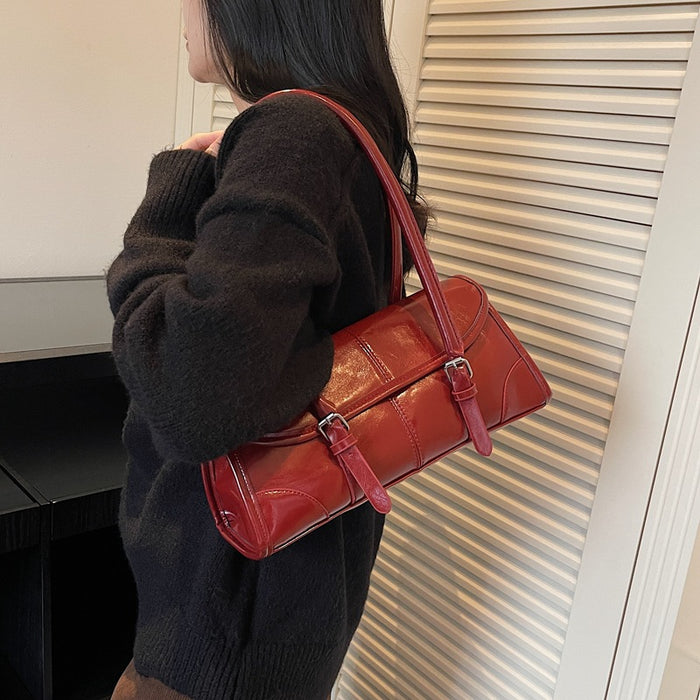 A person wearing a dark brown fuzzy sweater carries a glossy red shoulder bag with two front buckle straps, standing indoors against a white slatted door.