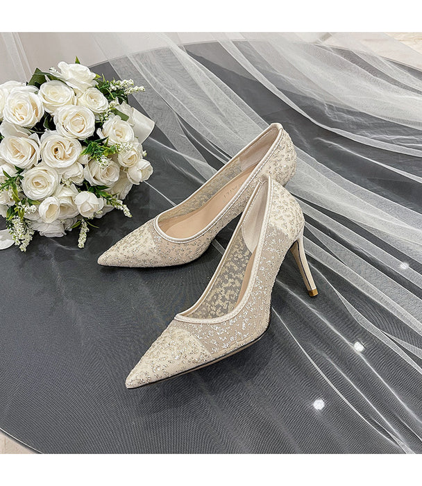 Pair of lace high-heeled shoes next to a bouquet of white roses on a gray surface.