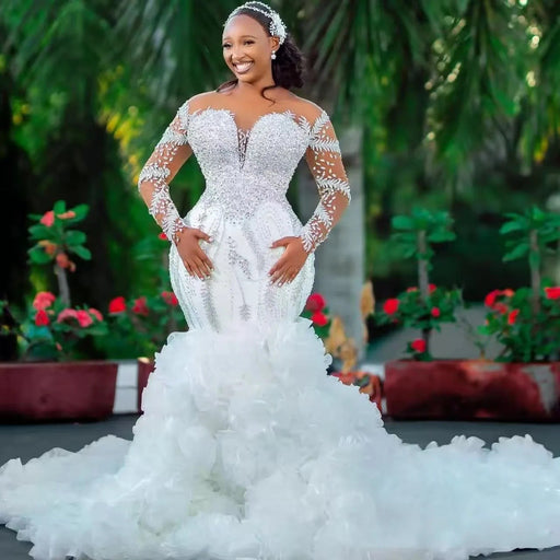 Woman in a white wedding dress with long sleeves and a train, standing outdoors with greenery in the background.
