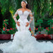 Woman in a white wedding dress with long sleeves and a train, standing outdoors with greenery in the background.