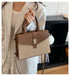 A woman in a white blazer holds a structured handbag with a woven beige body, brown leather trim and handles, and a gold-tone front clasp. The setting appears to be an indoor room with a dark shelf in the background.