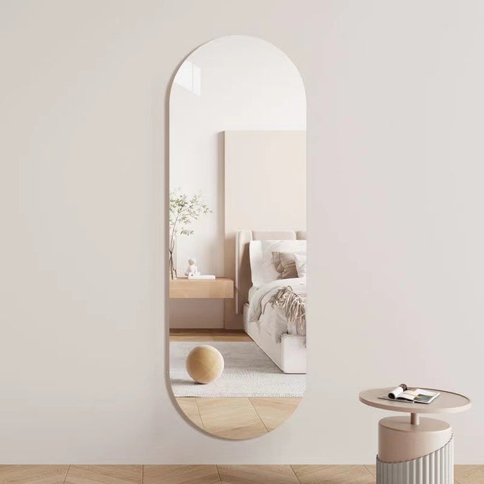 An arched full-length wall mirror with a slim frame, mounted on a light beige wall. The mirror reflects a softly styled bedroom interior with a bed, bedside table, and a round wooden sphere on the floor.