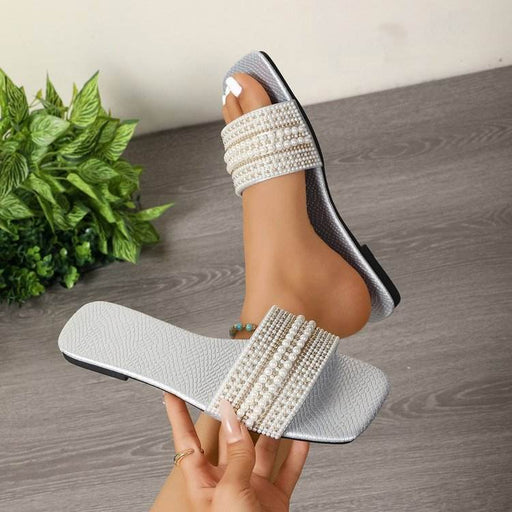 Pearl-embellished slide sandals being held on a wooden floor with a plant in the background