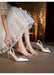 White high-heeled shoes with decorative elements worn by a person in a white lace dress.