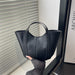 A black leather tote bag with dual top handles, held by a hand wearing a dark gray knit sleeve. The bag is positioned in front of a window with white blinds, next to a black frame and a book.