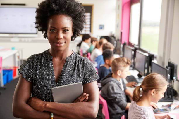 Teacher holding a tablet in a classroom with students working on computers