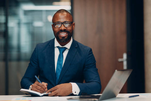 A smiling man in a navy blue suit, white dress shirt, and blue tie sits at a desk writing in a notebook with a pen, with an open laptop in front of him.
