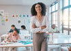 A smiling woman with curly hair stands in a bright classroom with children studying at desks. She is wearing a white long-sleeve blouse with a lace-trimmed V-neck and taupe trousers.
