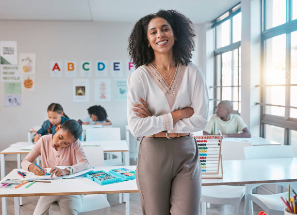 A smiling woman with curly hair stands in a bright classroom with children studying at desks. She is wearing a white long-sleeve blouse with a lace-trimmed V-neck and taupe trousers.