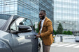 Man in a brown blazer getting into a car with modern buildings in the background