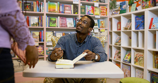 A man in a blue striped shirt sits at a white table in a bookshop, holding an open book. Bookshelves filled with colorful children's and adult books are visible in the background, and another person is partially visible on the left side of the frame.