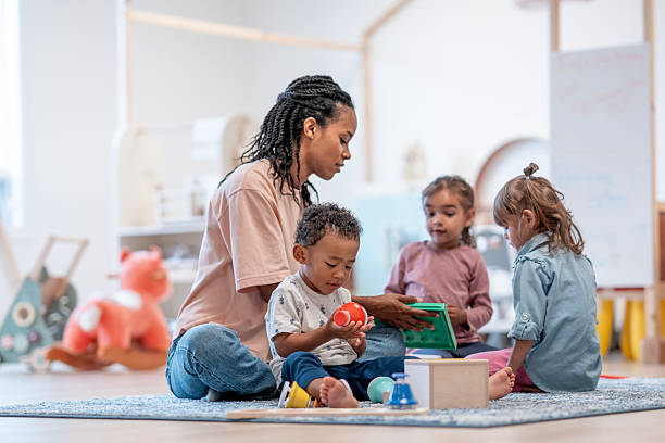 Woman playing with children on a rug in a room with toys.